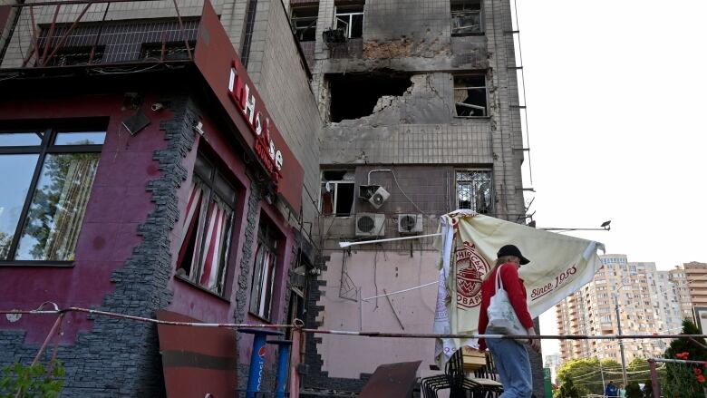 A person walks past a building with a large hole and other damage on it.