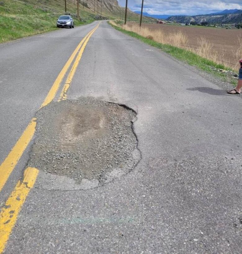 A large pothole in a sunny road with moutains and grass nearby.