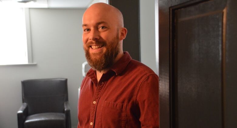 A smiling young bald man with a beard stands in an empty office