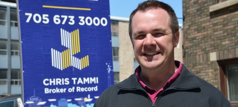 A man with short brown hair smiles for the camera in front of a for lease sign that has his name on it 