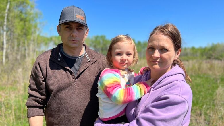 A man and a woman, the latter holding a three-year-old child, stand in a field.