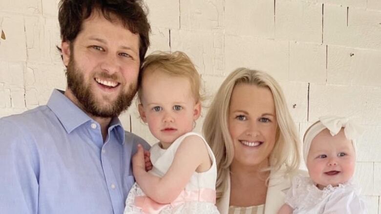 Smiling woman and man hold two very young children dressed in white.  