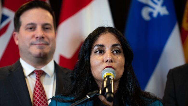 A South Asian woman is pictured speaking at a podium, with a man behind her.