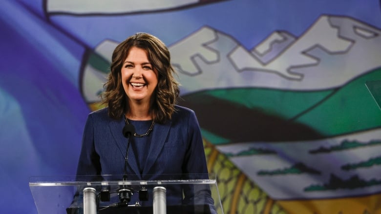 A woman smiles at a crowd from a podium with the Alberta flag behind her.