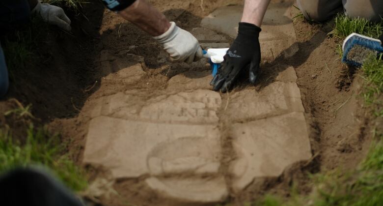 Two sets of hands are seen brushing earth from shards of a broken headstone lying flat in a freshly dug hole.