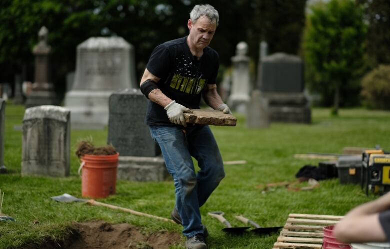 A man carries a large fragment of a broken headstone in a graveyard, as other headstones can be seen in the background.