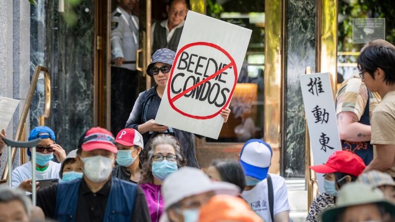 A group of people, some with masks, sit in front of a building. One woman holds a placard that reads 'Beedie Condos' with a cross sign on top of it.