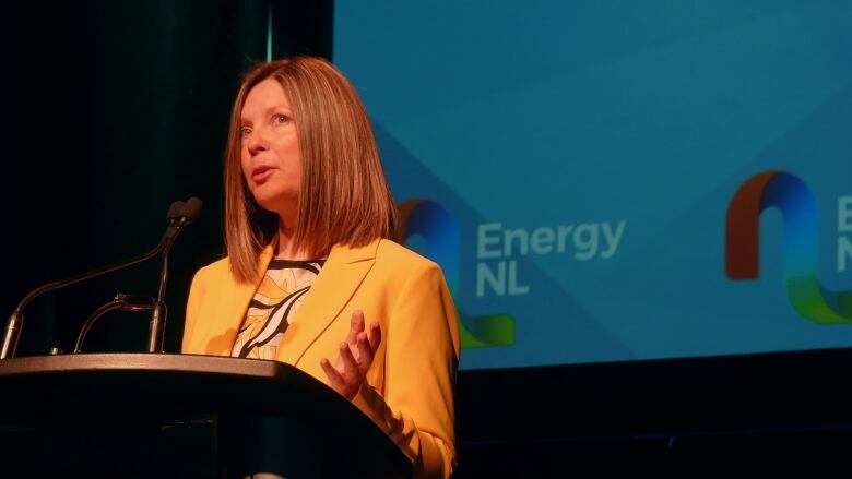 A woman with blonde-brown hair in a yellow blazer standing at a podium with the Energy NL logo behind her.