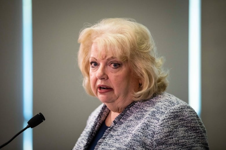 A woman with short blonde hair and dark eyeliner wears a grey textured blazer and stands at a white podium.