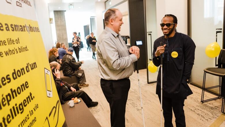 Two people holding white walking sticks chat and smile in a hallway.