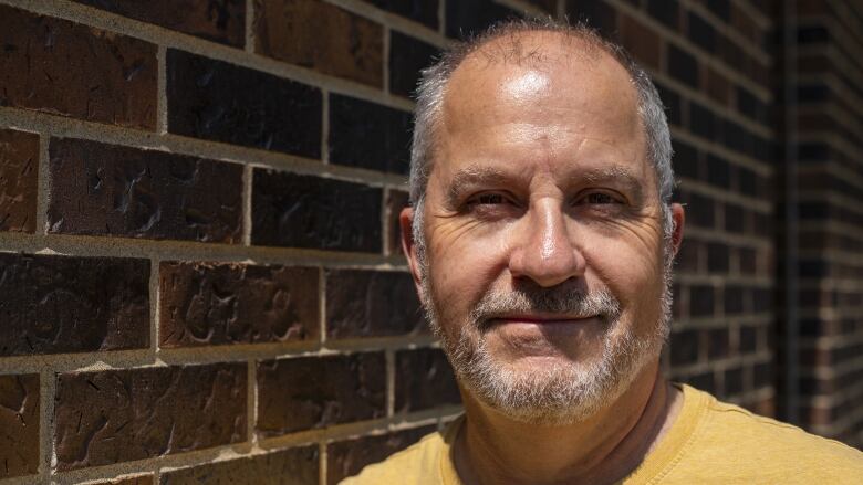 A man wears a yellow shirt while standing by a brick wall.