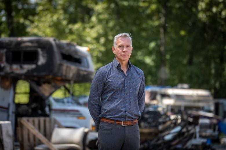 A well-dressed man in a clean open-necked blue shirt stands in front of old trailers.