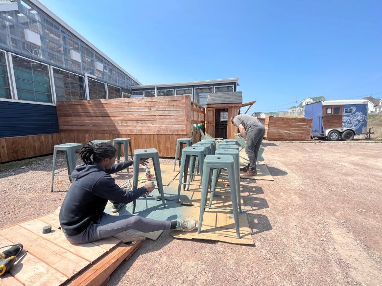 Two employees paint bar stools in the gravel parking lot.