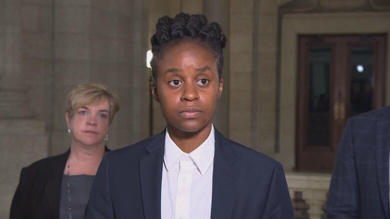 A person with black hair in a blue suit looks off camera, while a person with short blonde hair stands in the background.