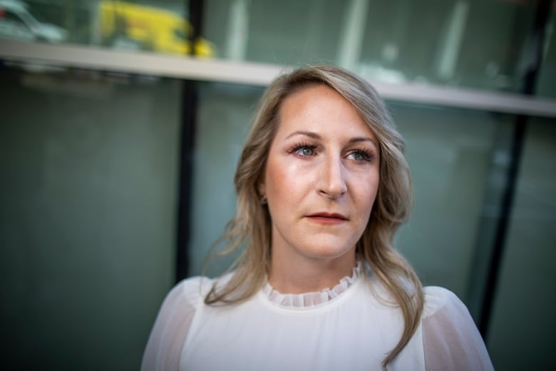 A woman with curled blonde hair wearing a white dress poses for a photo in front of a glass building.