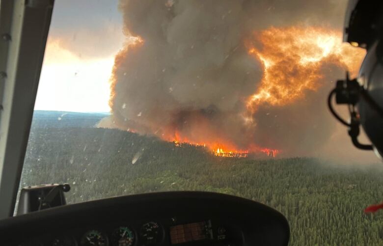 A large fire can be seen in a forest with foreground of a helmet and helicopter controls frame the shot.