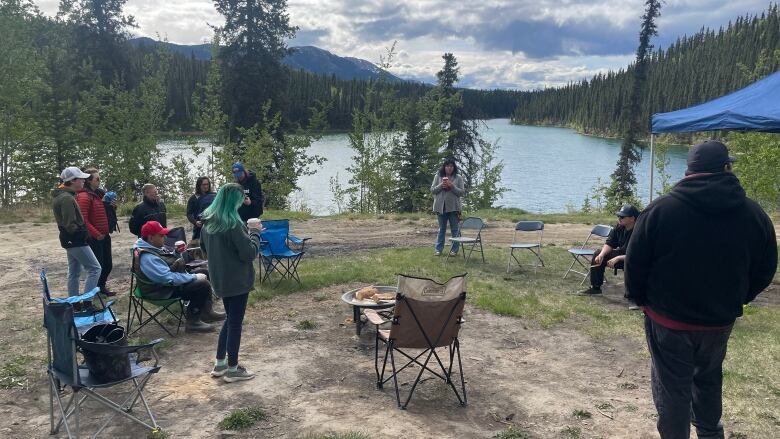 People stand around a fire with a lake and mountain range in the background.