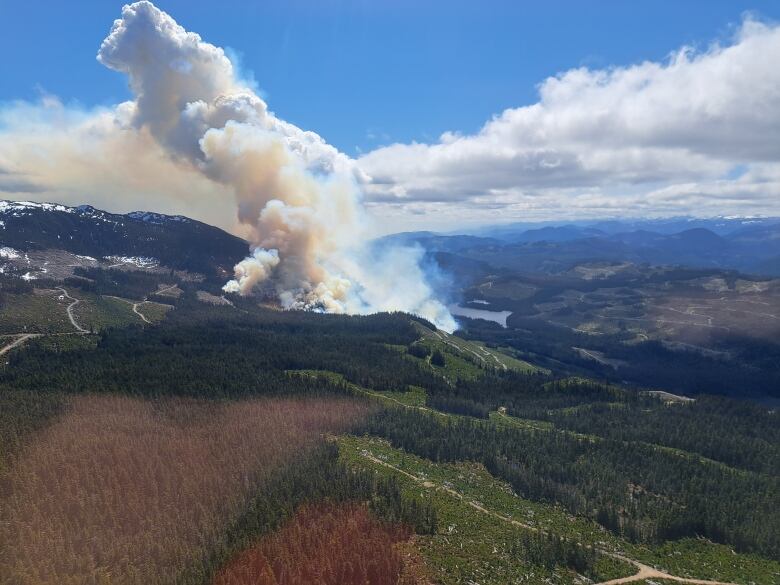 A large fire burns on green forested hills.