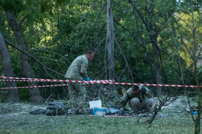 Two men in camouflage clothing work in a treed area set off by white and red caution tape.