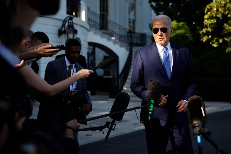 A silver-haired man in a suit and tie and wearing sunglasses stands in front of people and microphones outside.