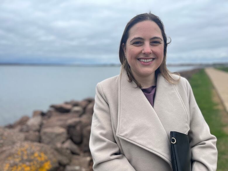 A woman stands against the backdrop of a river and boardwalk. She is smiling and wearing a beige coat. 