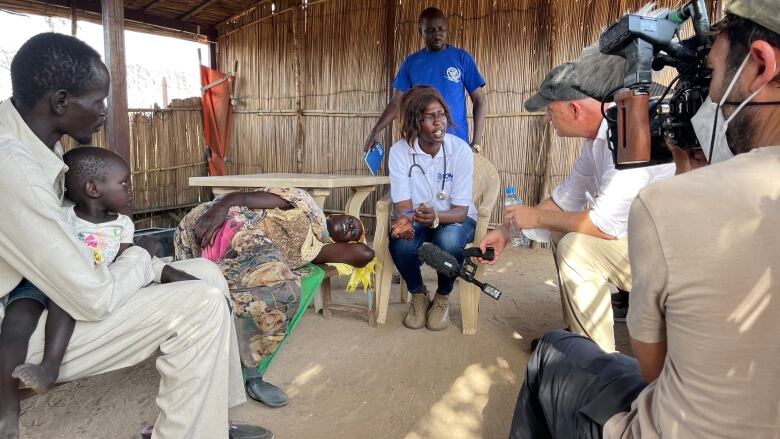 A group of people talk in a hut. One person is holding a camera and another is holding a microphone.