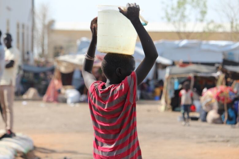 A child holds a bucket of water on their head.