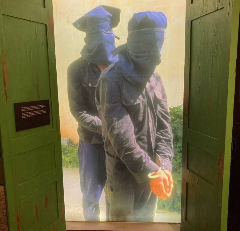 A damaged wooden door painted lime-green is open and shows two people with hoods over their heads, with their arms bound in front of them.