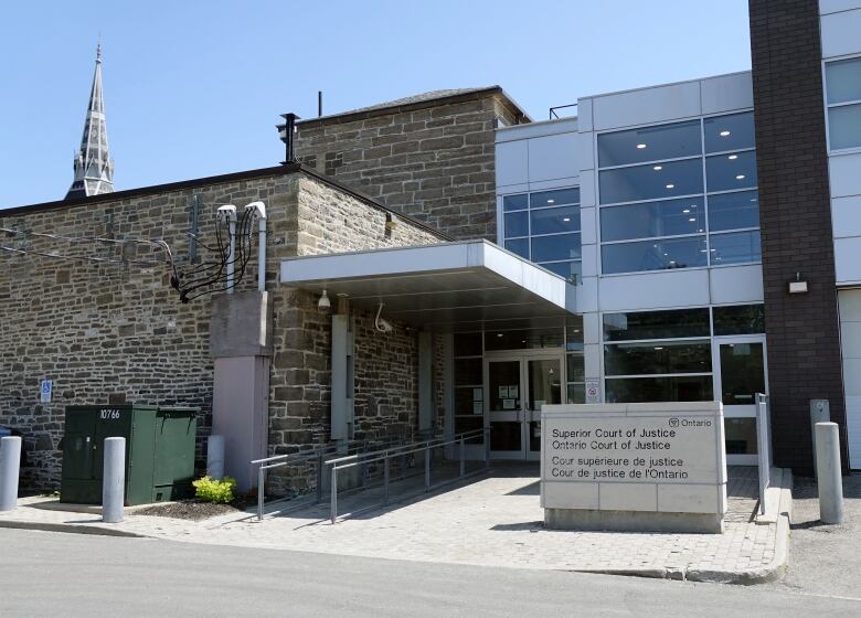 A grey stone and glass building with a sign identifying it as a courthouse out front is shown on a sunny day. A church steeple can be seen in the background.