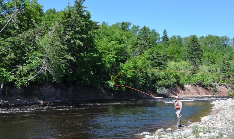 A man stands on the edge of a river, casting a fly fishing rod.