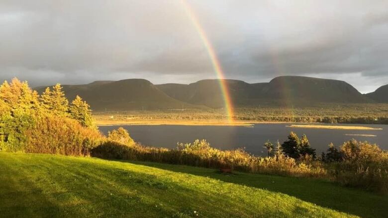 A double rainbow stretches across a river, with green grass in the foreground, and mountains and storm clouds in the background.