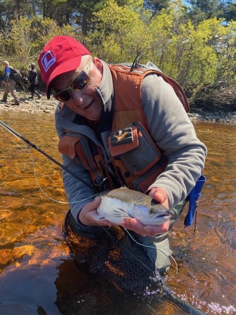 A man in a red cap stands in a river and holds out a salmon to the camera.