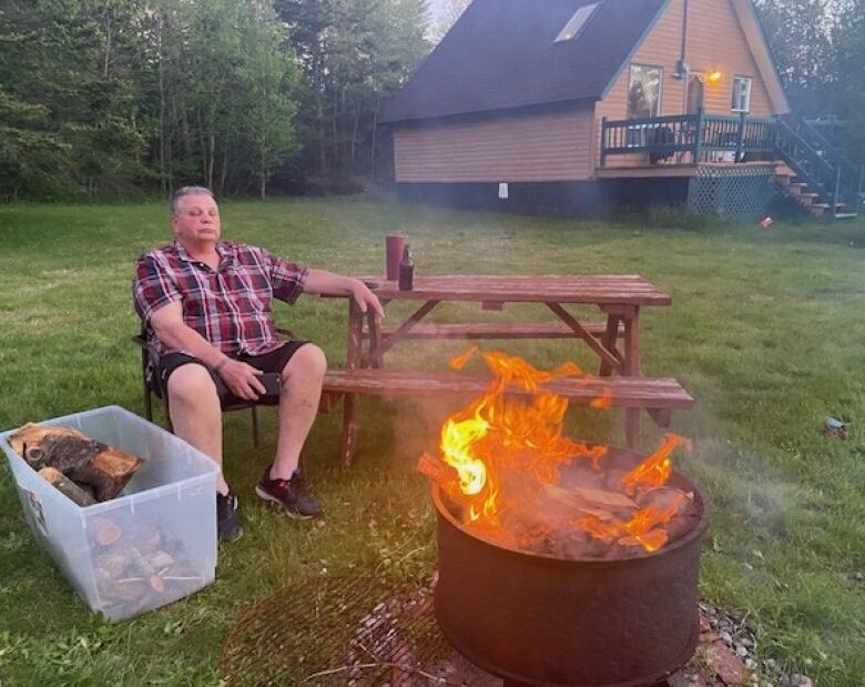 A man sits in front of a fire, beside a picnic table, with a cabin in the background.