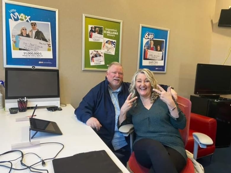 A woman with long blonde hair smiles at the camera, sitting in an office chair beside a large desk. Behind her, a man with short grey hair and a short grey beard also smiles in an office chair.