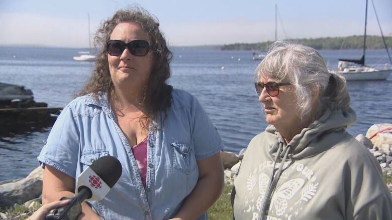 Two women are seen. The one on the left is taller, wit curly brown hair, wearing sunglasses and a light blue button up shirt. The one on the right is shorter, with grey hair and a grey hoodie.