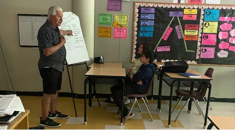 A man stands in front of two children in a classroom and points to a paper with words on it. 