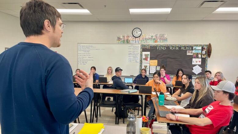 A teacher lectures a group of grade 12 students at Three Oaks Senior High.  Most students have laptops in front of them.  