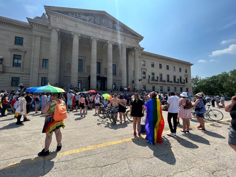 People stand facing the Manitoba Legislative Building. One person has a pride flag draped over their back.