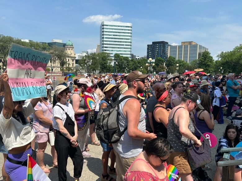 A crowd of people stand in front of the Manitoba Legislative Building. One person holds up a sign that says, 