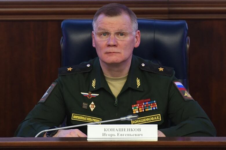 A man with glasses in a highly decorated military uniform sits in front of a microphone and a nameplate written in Cyrillic script.