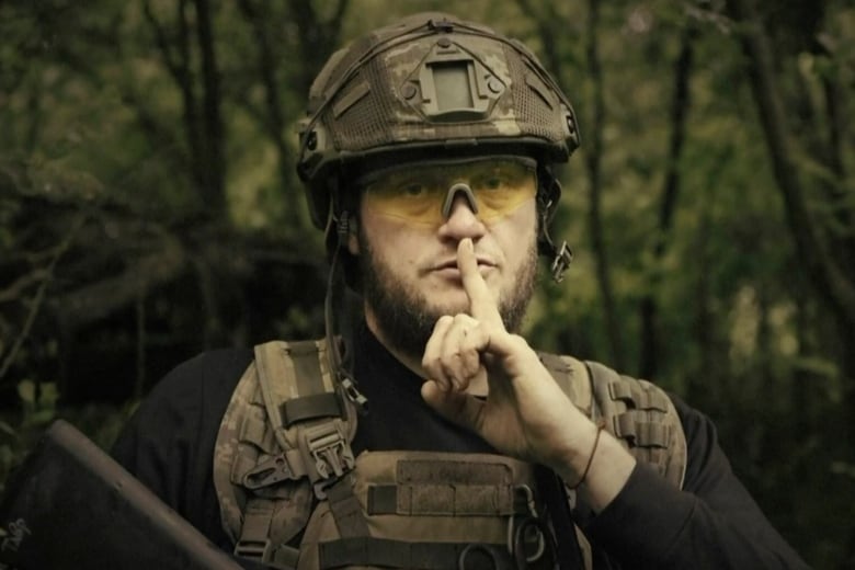 A bearded soldier wearing goggles and a camouflage helmet and vest presses his finger to his lips.