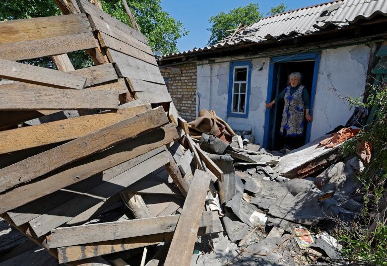 A woman is shown in the doorway of a house looking at wood and concrete debris in her yard.