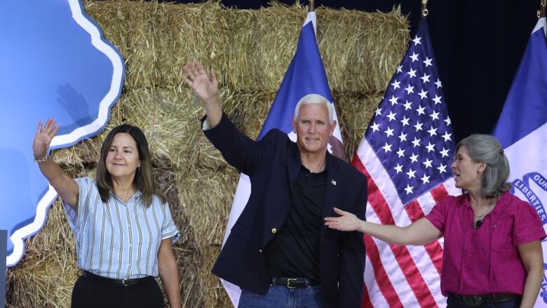 Two women and a man are shown onstage in front of a backdrop that includes bales of hay and an American flag.