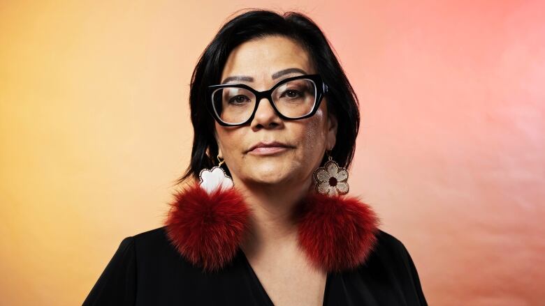 Head and shoulders image of a woman staring straight ahead, with dark hair and wearing dark glasses, a dark blouse and large red and white earrings.