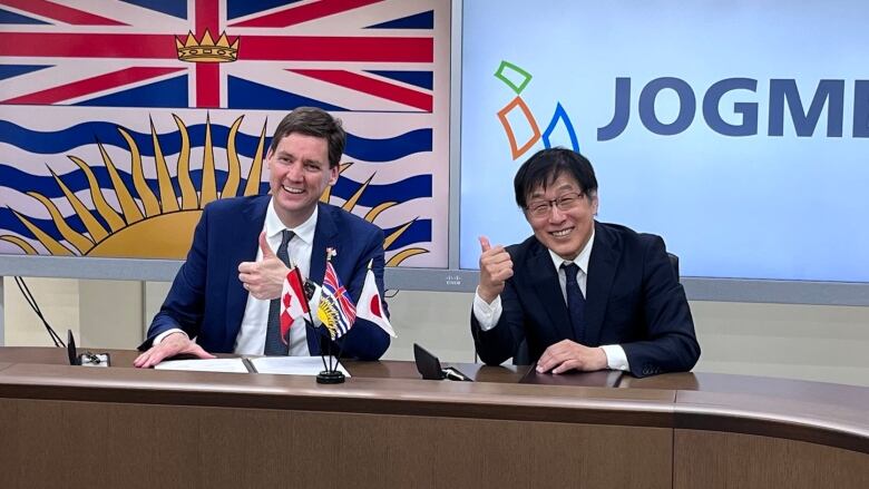 A smiling man in a suit and tie framed by the B.C. flag behind him gives a thumbs-up as he sits next to another smiling man in a suit and tie, also giving the thumbs-up with a logo behind him on the wall over his left shoulder that says JOGMEC.