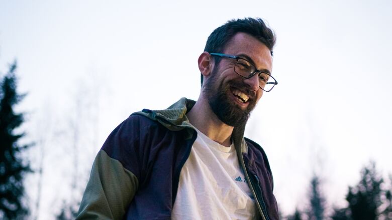 A man with a beard and glasses stands outside grinning from ear to ear. Conifer trees line the horizon behind him. 