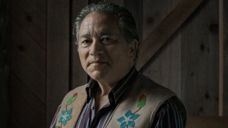 A portrait of an Indigenous man with grey hair, wearing a vest and a collared shirt looking into the camera.