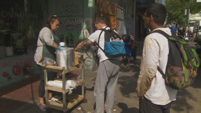 Rachel Chen holds a ladle by a crock pot full of soup on a cart. She is stationed in front of the Ovaltine Cafe, and two people are lined up in front of her stand. 