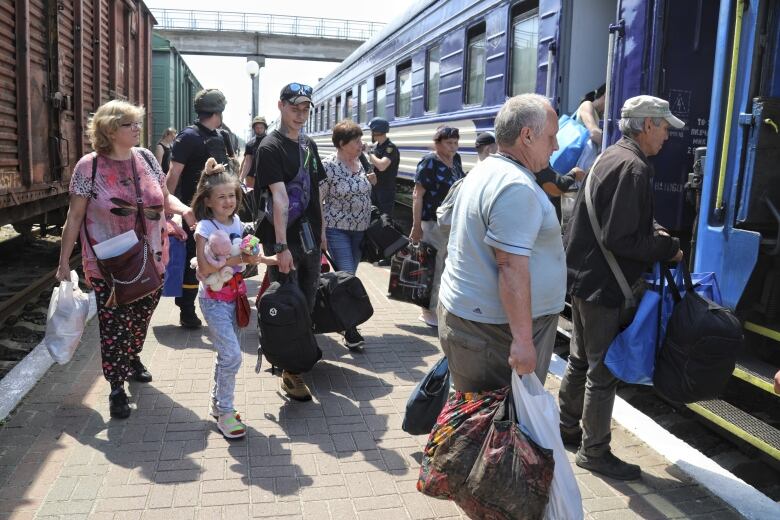 Men, women and a young girl holding several stuffed animals in her arms are shown on the platform of a station, about to board a train.