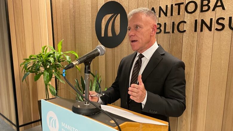 A man in short blonde hair and a dark suit stands at a podium in front of a wood panel wall.